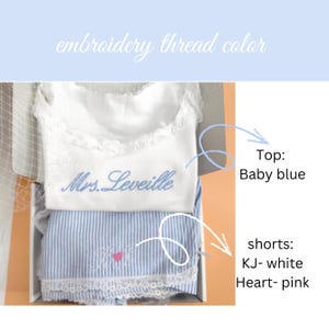 May include: A white lace-trimmed top embroidered with the text Mrs. Leveille in baby blue thread, paired with blue and white striped shorts featuring white embroidery and a pink heart. The text embroidery thread color is visible.