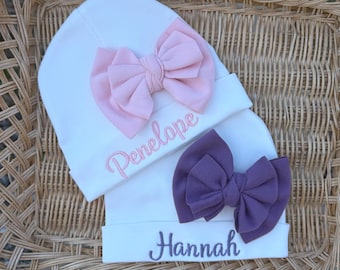 May include: White baby hats with embroidered names and large fabric bows. One hat has a pink bow and the name "Penelope" embroidered in pink. The other hat has a purple bow and the name "Hannah" embroidered in purple.