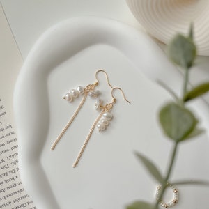 May include: A pair of gold dangle earrings featuring a delicate chain and small white pearls. The earrings have a simple and elegant design.