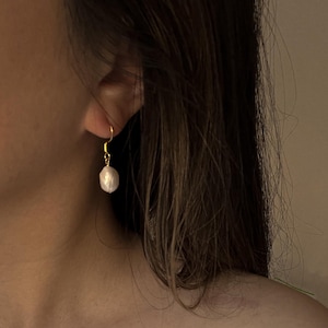 May include: A single gold earring featuring a white, irregularly shaped pearl. The earring hangs from an ear, with long brown hair in the background. The design is simple and elegant, showcasing the pearl's natural form.