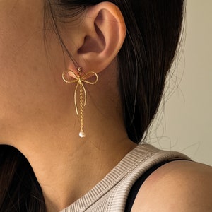 Puede incluir: Pendientes de lazo dorados con una perla que cuelga de la parte inferior del lazo.