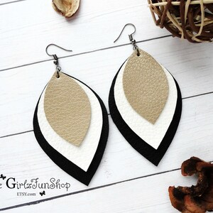 May include: A pair of layered leather earrings with black, white, and gold leaf shapes. The earrings are dangle style and have a silver hook.