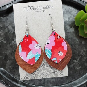 May include: A pair of dangle earrings with a red and pink floral pattern on a brown wooden base. The earrings are hanging from silver hooks.