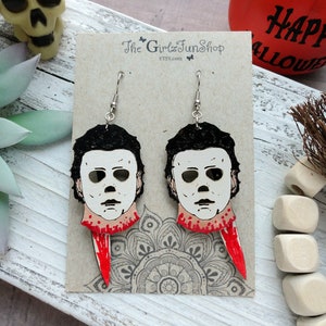 May include: A pair of Halloween-themed earrings featuring a white mask with black eyes and a red slash across the neck. The earrings are on a brown card with the text "The Girlz Fun Shop Etsy.com".