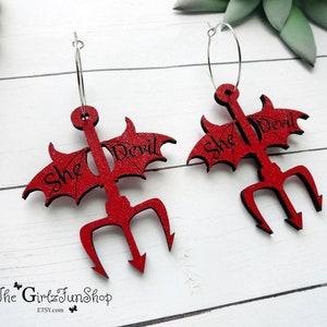 May include: A pair of red glitter earrings shaped like a devil's trident with wings. The earrings are made of wood and have the text "She Devil" on them.