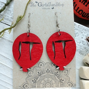 May include: Red wooden earrings shaped like a balloon with the word "IT" cut out. The earrings are hanging from silver hooks.