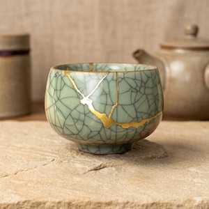 May include: A small, round, celadon-colored ceramic tea bowl with a cracked glaze pattern and gold-colored kintsugi repair. The bowl has a low, rounded shape and is likely used for tea ceremonies.