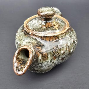 Puede incluir: Una tetera de cerámica con tapa y boquilla. La tetera tiene un aspecto moteado y jaspeado en tonos verdes, marrones y blancos. La tapa tiene un pequeño pomo y la boquilla es curva. La tetera está diseñada para preparar té.