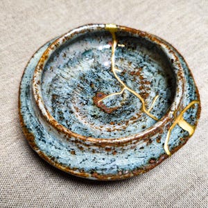 May include: A small, round ceramic dish featuring a mottled blue and brown glaze with a textured, crackled surface. The dish displays a prominent repair line filled with gold-colored lacquer, extending from the rim down into the center basin.