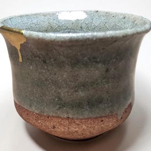 May include: A ceramic cup with a flared rim, featuring a textured gray-green glaze on the upper portion and an exposed, rough terracotta-colored base. A single gold-colored metallic accent is applied to the top edge of the rim.