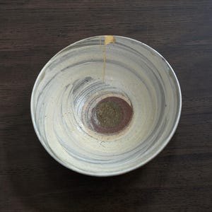 May include: A ceramic bowl with a cream-colored exterior and a spiral design in shades of gray and brown. The center features a dark brown and green circle. A gold-colored line runs down the side of the bowl. The bowl is on a dark brown surface.