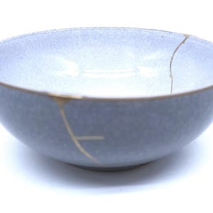 May include: A light blue ceramic bowl with a gold rim and gold-filled cracks, showcasing the Japanese art of Kintsugi. The bowl has a smooth, matte finish and a simple, elegant design.