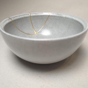 May include: A light gray ceramic bowl with a smooth, matte finish. The interior of the bowl features a distinct, thin, Y-shaped gold line that extends from the rim down toward the center, highlighting a repair or decorative accent on the surface.