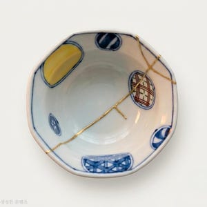 May include: A white ceramic bowl with an octagonal rim, featuring blue and yellow hand-painted geometric and circular patterns. A prominent gold-colored repair line runs across the interior surface, highlighting the kintsugi technique.