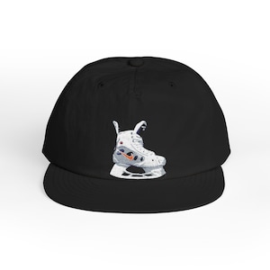 May include: A black baseball cap featuring an embroidered graphic of a white ice skate on the front panel. The skate design includes a small orange swoosh logo, grey accents, and white laces, centred on the crown above the flat brim.