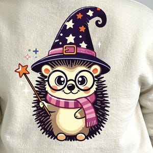 May include: A cream-colored sweatshirt featuring a cartoon hedgehog wearing a wizard hat with stars, glasses, and a striped scarf. The hedgehog holds a star-shaped wand, with magical sparkles around it. The design is whimsical and playful.