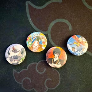 May include: Four circular pin-back buttons featuring animated characters from Avatar: The Last Airbender. One button shows Appa, another features Aang and friends, one displays Zuko with fire, and the final button depicts Aang, Katara, and Sokka.