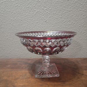 May include: A ruby red glass pedestal bowl with a square base and a decorative, diamond-cut pattern. The bowl has a clear, textured stem and a scalloped rim. The bowl is likely used for serving fruit or desserts.