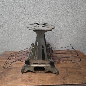 Puede incluir: Una tostadora de metal antigua con un diseño floral en la parte superior y rejillas de alambre. La tostadora tiene un acabado plateado con perillas negras y está colocada sobre una superficie de madera. Un electrodoméstico de cocina vintage.