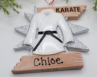May include: White karate uniform Christmas ornament with a black belt, a silver starburst background, and a wooden sign that reads "KARATE" and "Chloe".