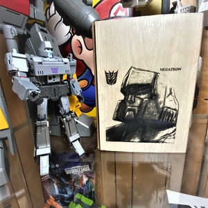 May include: A wooden box with a black and white drawing of Megatron, a Transformers character, on the front. The box is labeled "Megatron".