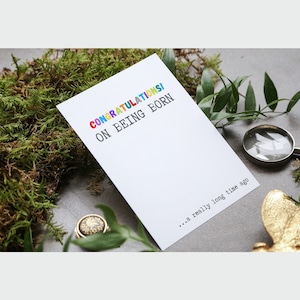 May include: A white card with the text "CONGRATULATIONS! ON BEING BORN... a really long time ago" in colorful letters. The card is sitting on a bed of green moss.