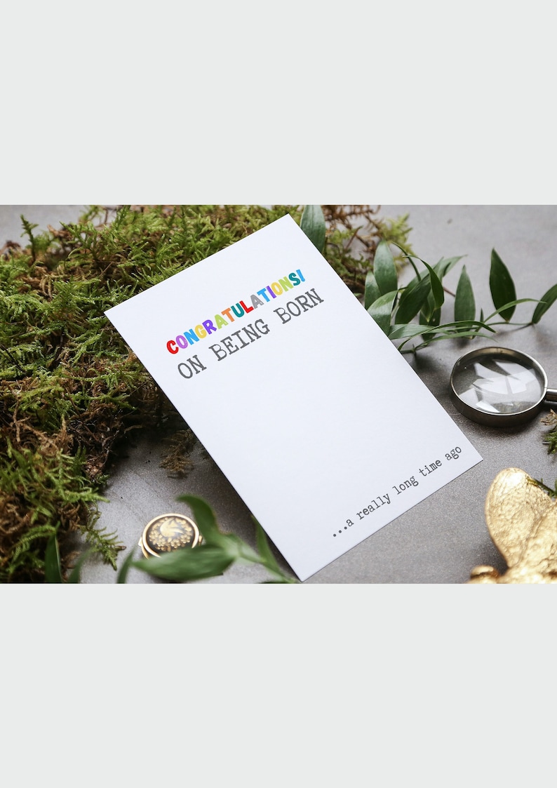 May include: A white card with the text "CONGRATULATIONS! ON BEING BORN... a really long time ago" in colorful letters. The card is sitting on a bed of green moss.