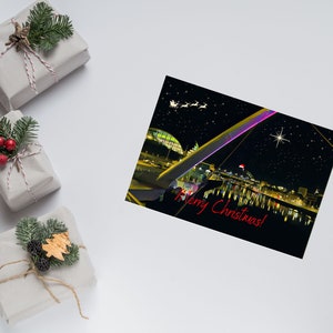 Può includere: Immagine a tema natalizio con regali confezionati e un biglietto di auguri. Il biglietto mostra un paesaggio notturno con un ponte, la scritta "Merry Christmas!" e una slitta di Babbo Natale. I regali sono avvolti in carta marrone con verde festivo.