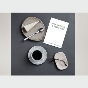 May include: A white card with the text "CONGRATULATIONS! ON EEING BORN... a really long time ago" in rainbow colors. The card is on a gray surface with a cup of coffee, a plate with a slice of cheesecake, a spoon, and a pair of glasses on a small white dish.