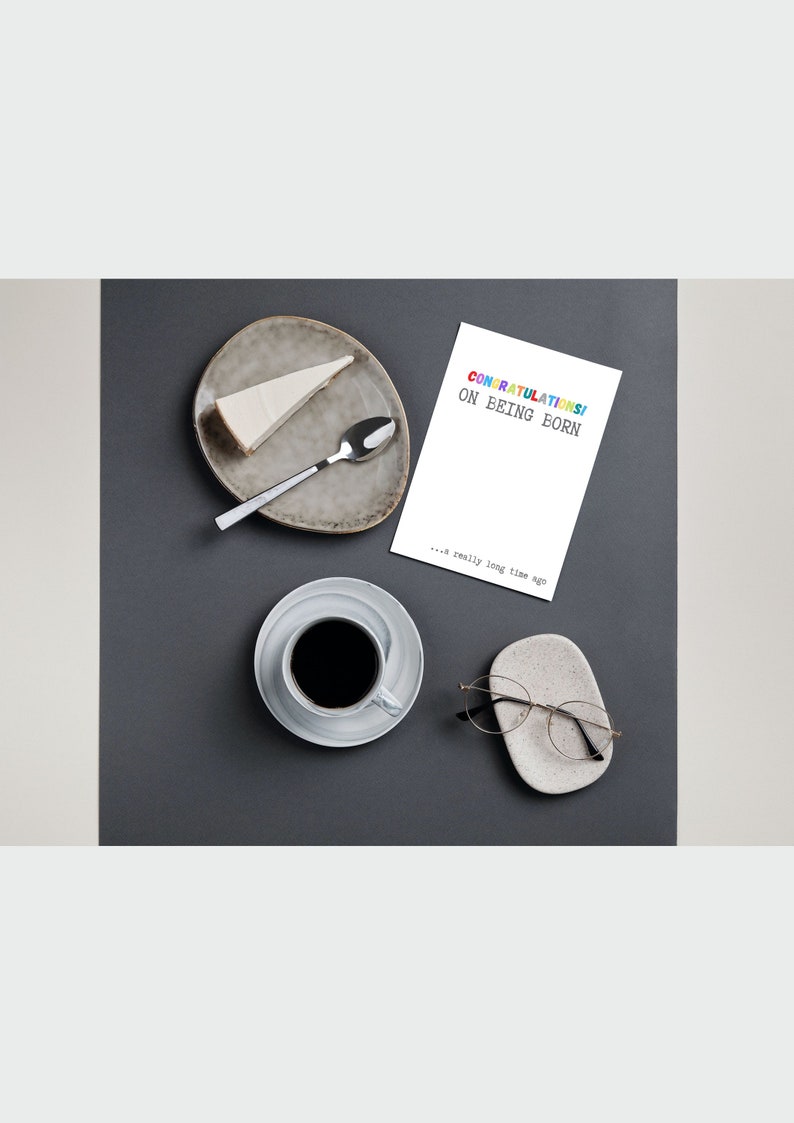 May include: A white card with the text "CONGRATULATIONS! ON EEING BORN... a really long time ago" in rainbow colors. The card is on a gray surface with a cup of coffee, a plate with a slice of cheesecake, a spoon, and a pair of glasses on a small white dish.