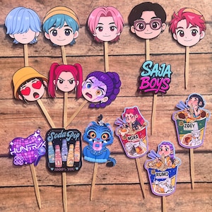 May include: A collection of colorful cartoon character cutouts on wooden sticks. The characters include faces with various hairstyles and expressions, along with text that says "SAJA BOYS", "HUNTRX", and "Soda Pop". There are also images of ramen bowls and a blue tiger.
