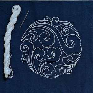 Op de afbeelding: Close-up van een marineblauwe stof met een wit geborduurd cirkelvormig ontwerp met wervelende wolkenachtige patronen. Een witte draadspoel en naald bevinden zich naast het ontwerp.
