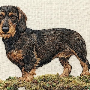 Puede incluir: Bordado detallado de un perro salchicha, con tonos marrones y negros. El perro está sobre una superficie herbosa texturizada, sobre un fondo neutro. La obra destaca los rasgos distintivos del perro.