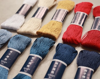 Puede incluir: Una colección de diez madejas de hilo de bordar dispuestas en dos filas. Los colores incluyen blanco, crema, amarillo, rojo y varios tonos de azul. Cada madeja está asegurada con una banda de papel azul oscuro con texto blanco y caracteres.