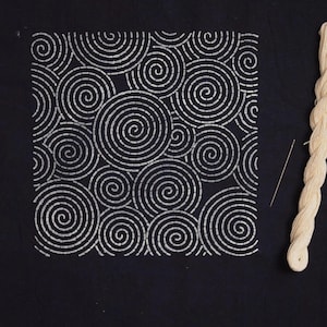 Könnte beinhalten: Ein schwarzes quadratisches Stoffstück mit einem weißen Spiralmuster, einer Nadel und einem cremefarbenen Fadenbündel. Das Spiraldesign besteht aus kleinen, eng beieinander liegenden Stichen, die einen texturierten Effekt erzeugen.