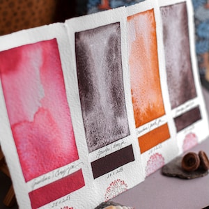 Könnte beinhalten: Vier Aquarell-Farbmuster auf weißem Papier, die verschiedene Schattierungen von Rot, Braun, Orange und Schwarz zeigen. Die Farbmuster sind mit den Namen der Farben und dem Datum ihrer Entstehung beschriftet.