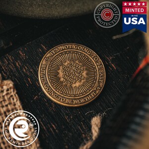 May include: A bronze-coloured coin with the text "It's not going to be easy, but it's going to be worth it." and "Never forget the power you have inside you." The coin has a sunburst design with a starburst around the edge.