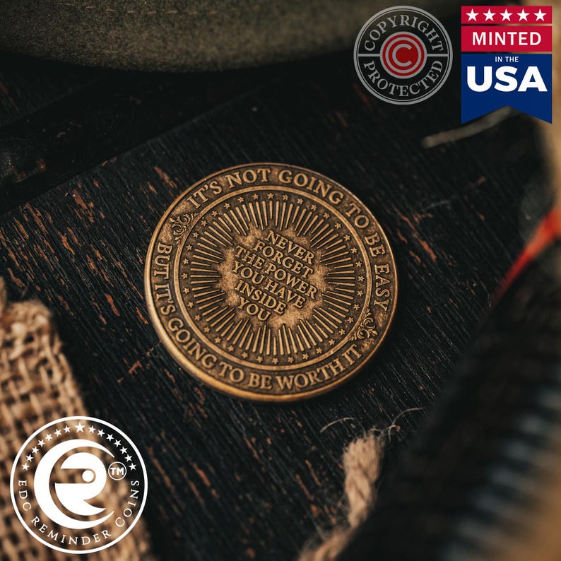 May include: A bronze-coloured coin with the text "It's not going to be easy, but it's going to be worth it." and "Never forget the power you have inside you." The coin has a sunburst design with a starburst around the edge.