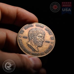 May include: A round bronze-toned metal coin featuring a relief portrait of Seneca in the center. The text reads WE SUFFER MORE OFTEN IN IMAGINATION THAN IN REALITY, with 4 BCE and 65 CE flanking the portrait and the name SENECA below it.