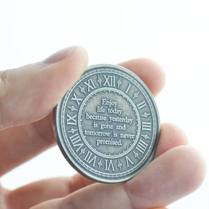 May include: A silver-toned round coin with a clock face and the inscription "Enjoy life today because yesterday is gone and tomorrow is never promised."