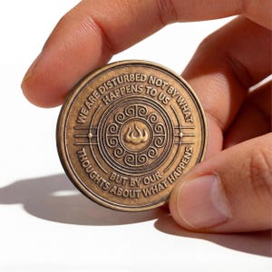 May include: A round bronze-colored metal coin featuring an embossed design with a central flame-like symbol surrounded by a spiral pattern. The text reads: WE ARE DISTURBED NOT BY WHAT HAPPENS TO US BUT BY OUR THOUGHTS ABOUT WHAT HAPPENS.
