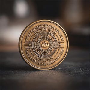 May include: A round bronze-toned metal coin featuring an embossed design with a central flame-like symbol surrounded by a circular pattern of swirls. The text reads: WE ARE DISTURBED NOT BY WHAT HAPPENS TO US BUT BY OUR THOUGHTS ABOUT WHAT HAPPENS.