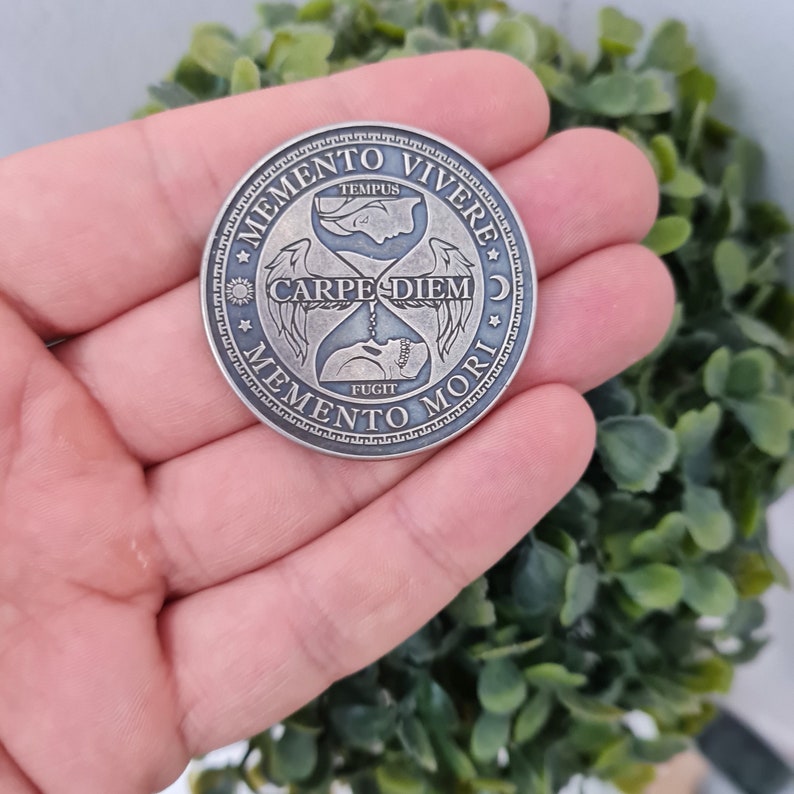 May include: A silver-toned coin with an hourglass design and the Latin phrase "Memento Vivere Carpe Diem Memento Mori" around the edge. The hourglass is surrounded by wings and stars.