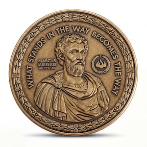 May include: A circular bronze-toned commemorative coin featuring a relief portrait of Marcus Aurelius. The text reads WHAT STANDS IN THE WAY BECOMES THE WAY, MARCUS AURELIUS AD 121-180, and STOIC with a flame icon. A laurel wreath border surrounds the design.