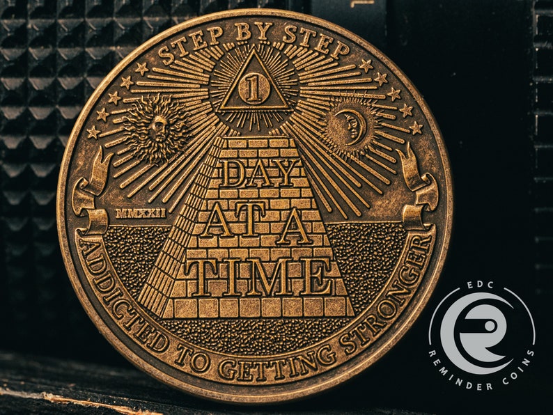 May include: A gold-toned metal coin with an intricate design featuring a pyramid, an all-seeing eye, and the text "Step by Step Day at a Time Addicted to Getting Stronger".