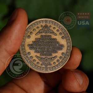 May include: A circular bronze-toned metal coin featuring an embossed border of stars and laurel leaves. The center displays the text: IT IS IN THE STRUGGLE THAT WE DISCOVER OUR TRUE POTENTIAL, FOR WHAT STAYS IN THE WAY SHAPES WHO WE BECOME.