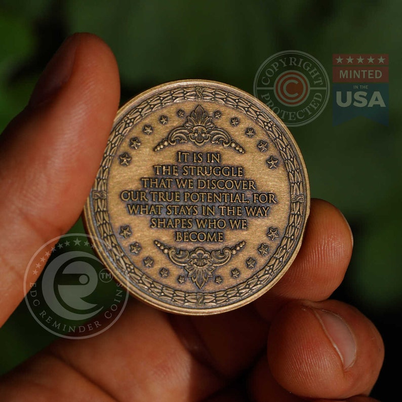 May include: A circular bronze-toned metal coin featuring an embossed border of stars and laurel leaves. The center displays the text: IT IS IN THE STRUGGLE THAT WE DISCOVER OUR TRUE POTENTIAL, FOR WHAT STAYS IN THE WAY SHAPES WHO WE BECOME.