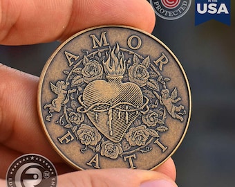 May include: A circular bronze-toned metal coin featuring an embossed design of a flaming heart encircled by roses and two cherubs. The text AMOR FATI is inscribed along the border. The coin has a weathered finish with dark recessed details.