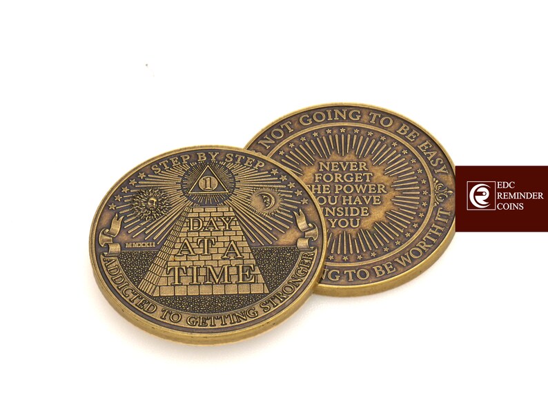 May include: A gold-toned metal coin with the words "Step by Step" and "Addicted to Getting Stronger" on the front. The back of the coin has the words "Not Going to Be Easy" and "Never Forget the Power You Have Inside You" on it.