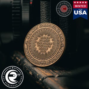 May include: A bronze-coloured coin with the inscription "It's not going to be easy, but it's going to be worth it." The coin also features the words "Never forget the power you have inside you."